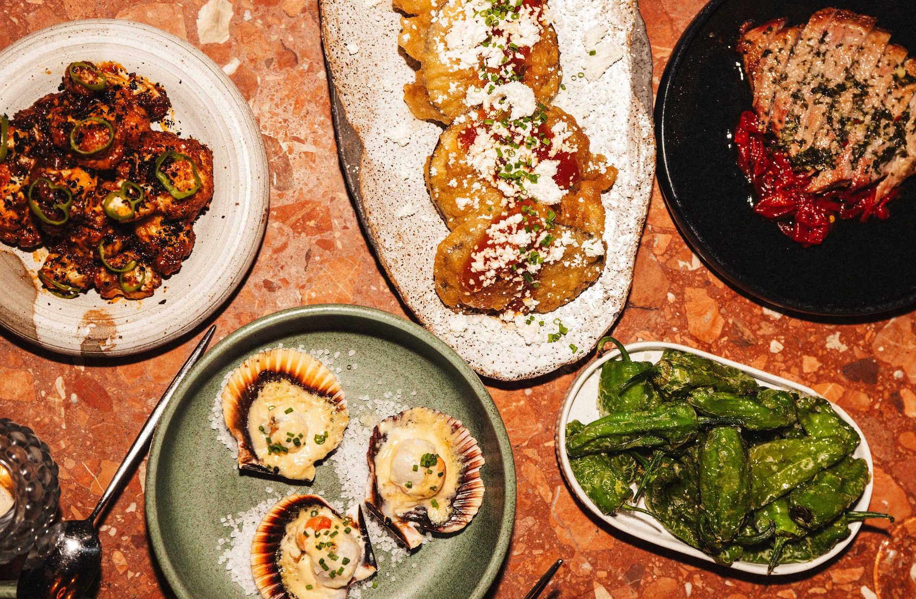 Sex tapasrätter ovanifrån på marmorbord – blomkål, aubergine, presa, kammusslor och padrón